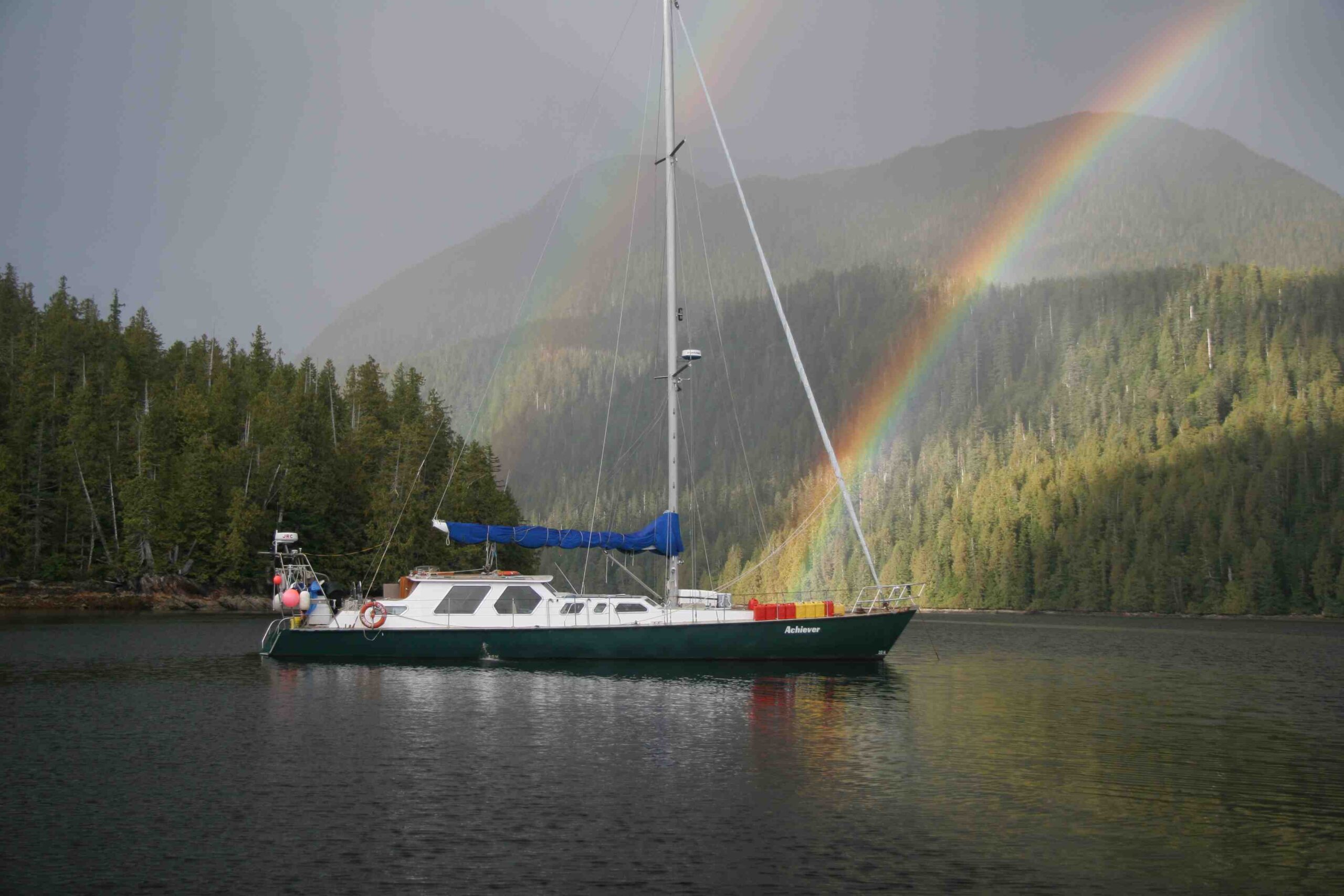 The Achiever, a 60-foot sailing vessel, sits on the water, while forested hills rise in the background. From the water, a double rainbow rises - the brighter rainbow seems to come from the bow of the boat.