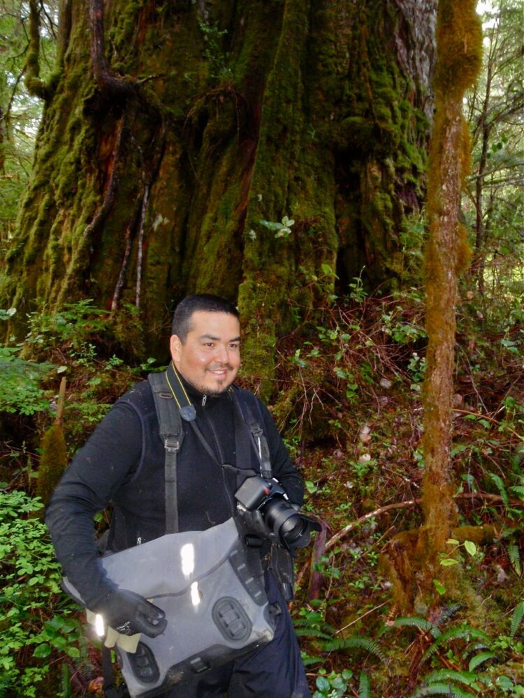 A man with a camera in front of a large tree.