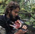 A man (Chris) holds a partially eaten fish in the forest.