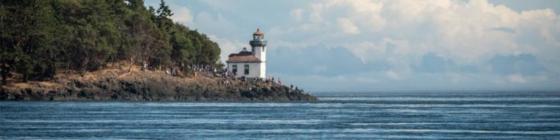 J50 swims toward San Juan Island: a group of people stand by the lighthouse waiting and watching.