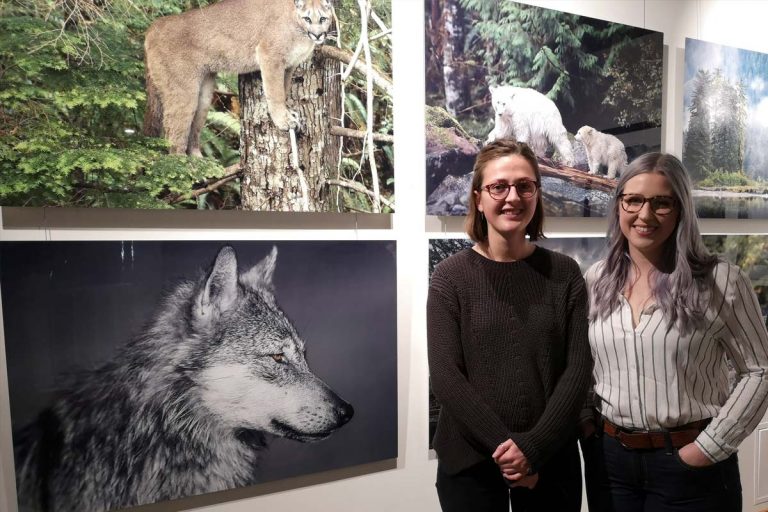 Two women stand in front of the One Shot for Coastal Carnivores exhibit at the Robert Bateman Centre.