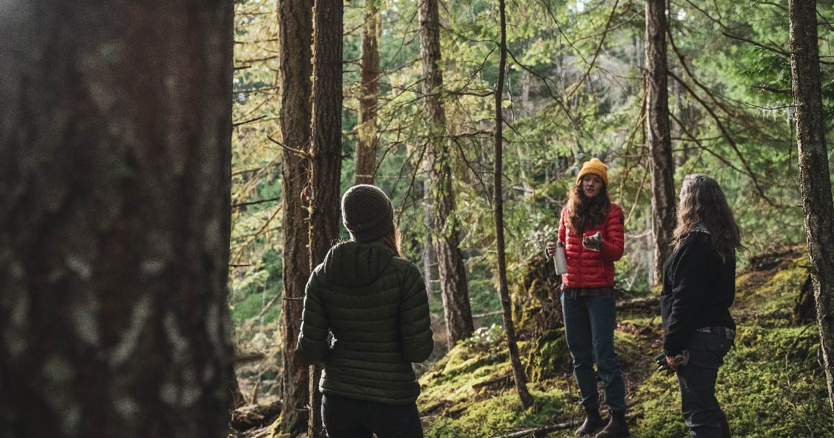 Three women standing 6 feet apart talking to each other in a Coastal Douglas-fir forest