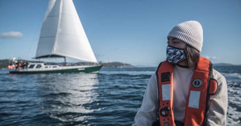 Young woman wearing a mask sitting on a boat with a sail boat in the background on a sunny day.