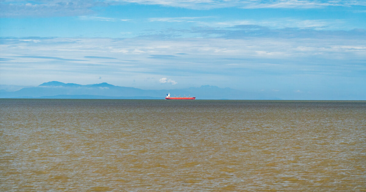 Landscape image with the estuary in the foreground, mountains in the background and a tanker centered over the water. 