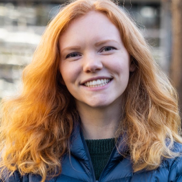 Raincoast Fellow and Masters of Science student Monica Short stands looking at the camera, with shoulder-length, wavy auburn hair, wearing a blue Rab puffy jacket and black knit pullover underneath.