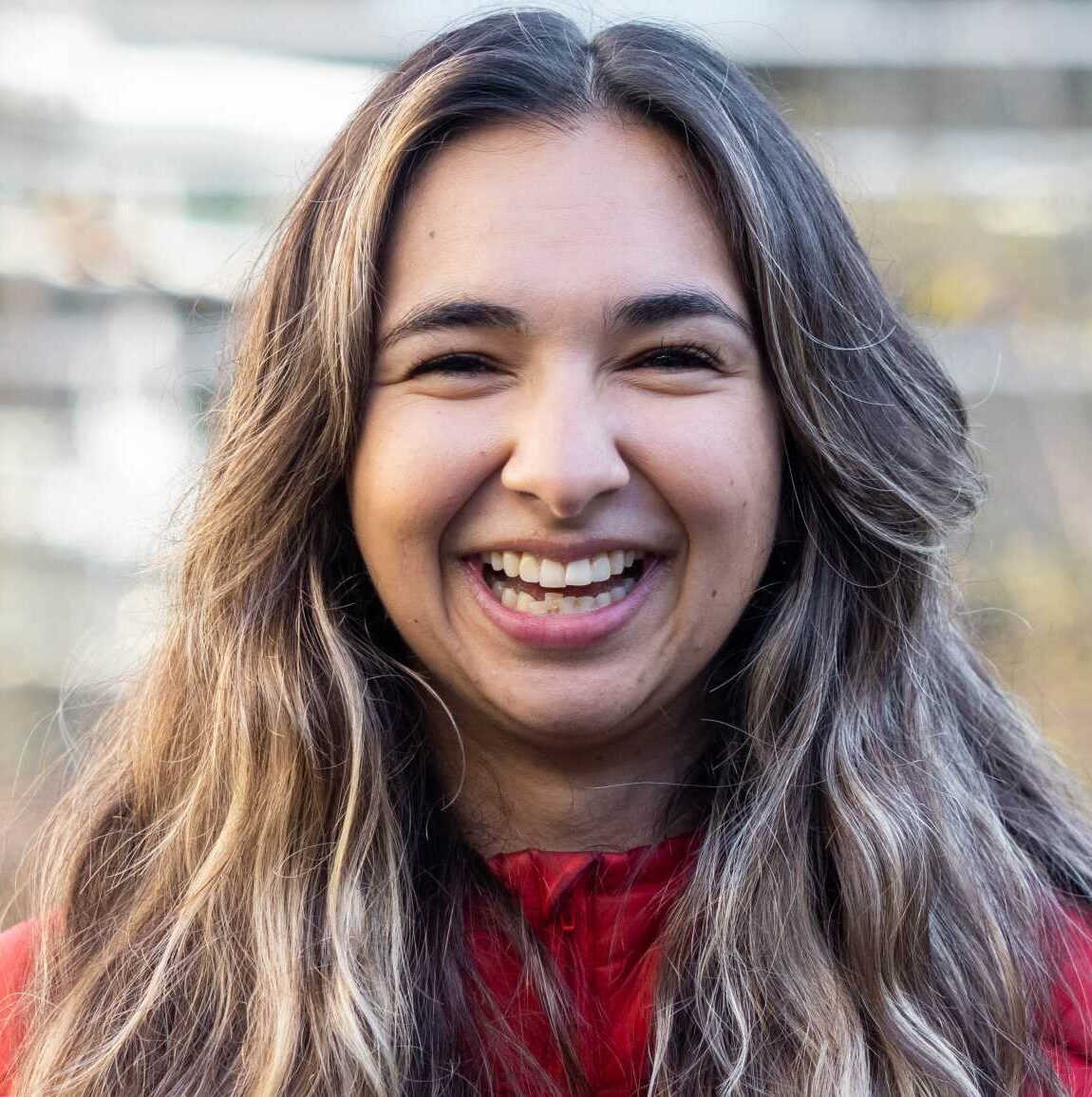 Persia Khan, Research Associate and Liaison for Raincoast, looks straight at the camera with a giant grint, showing off her teeth and crinkling her eyes. Her hair is a wavy mix of blonde and brown, settling over her shoulders while wearing a red puffy jacket.