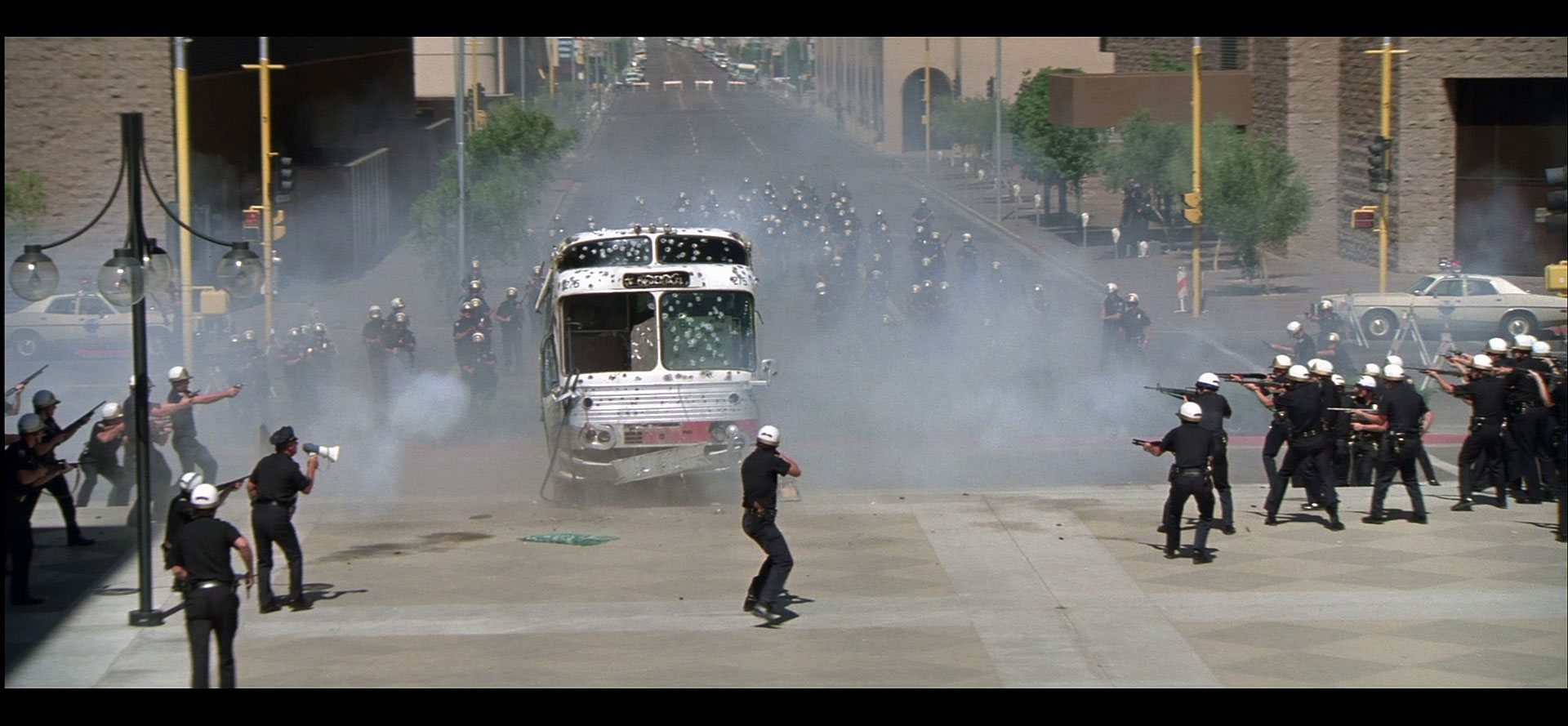 A bus riddled with bullet holes, surrounded by smoke, comes to a stop in the middle of hundreds of police with rifles shooting and shooting.