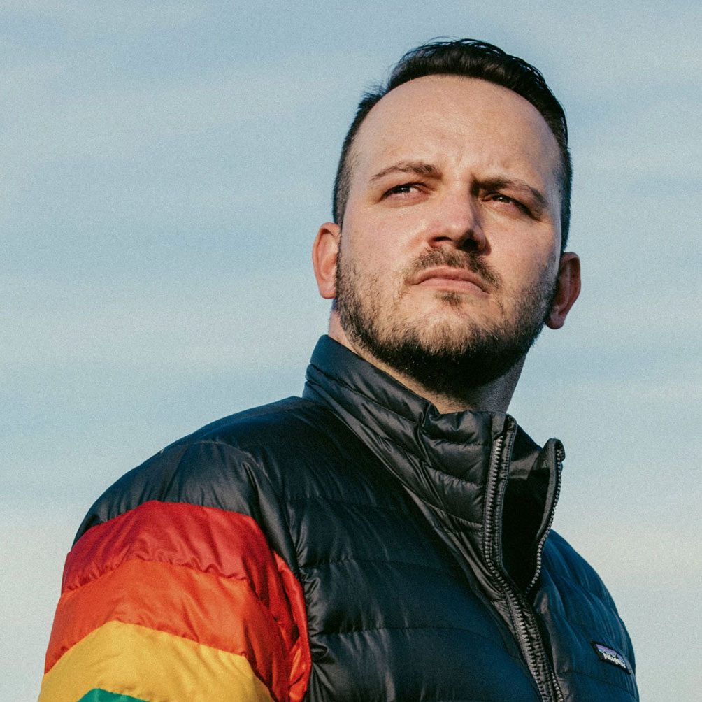 A portrait of Denis wearing a patagonia puffy coat with a rainbow on the arm.