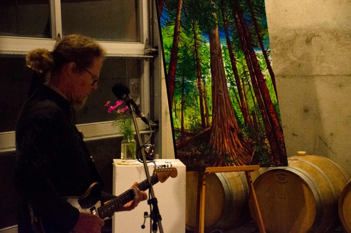 Man with a guitar singing next to a painting of a tree.