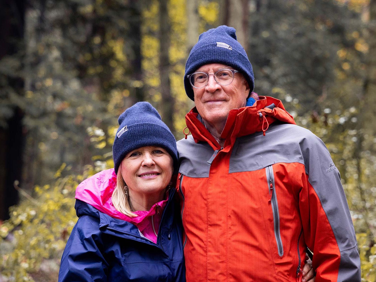 Jane and Ross Woodland smiling in a forest wearing blue Raincoast toques.