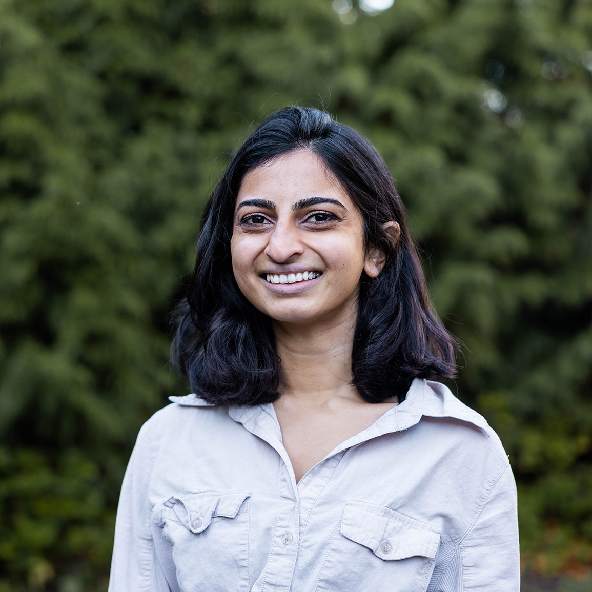 Raincoast Fellow and Masters of Science student Ishana Shukla looks at the camera, her hair is black and shoulder-length, wearing a light grey cotton button-up shirt.