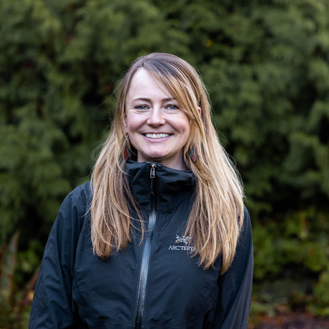 Kate Field, a team member of Raincoast, standing against a blurred backdrop of green cedar trees, looking at the camera smiling and wearing a black jacket that constrasts with her bright eyes and blonde hair.