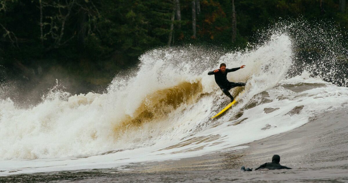 A surfer rips at the top of a wave on the central coast of BC with another surfer in the water in the foreground.