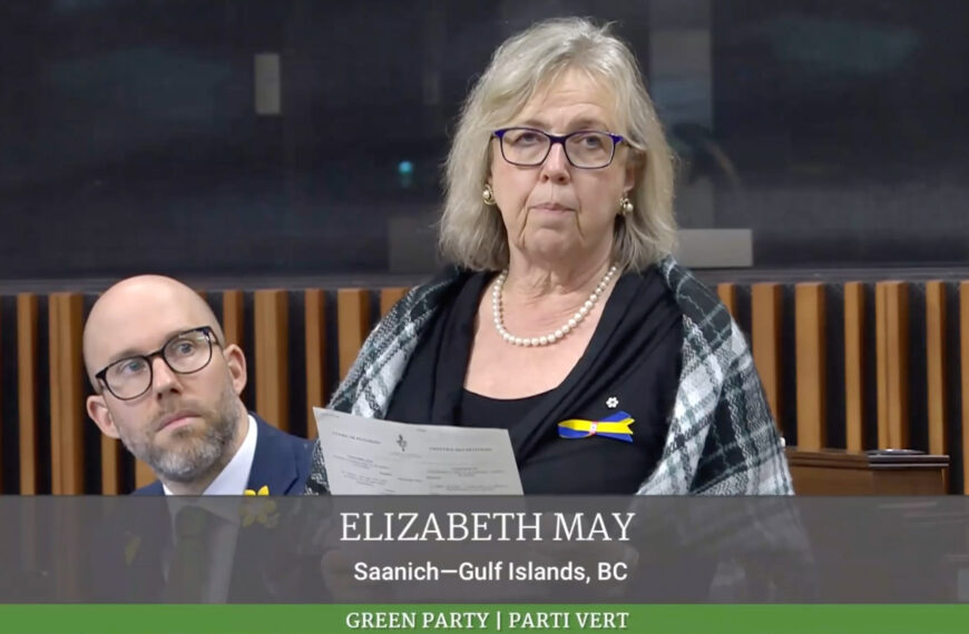Elizabeth May with a bald man wearing glasses behind her, holding a paper and looking concerned in a government building.