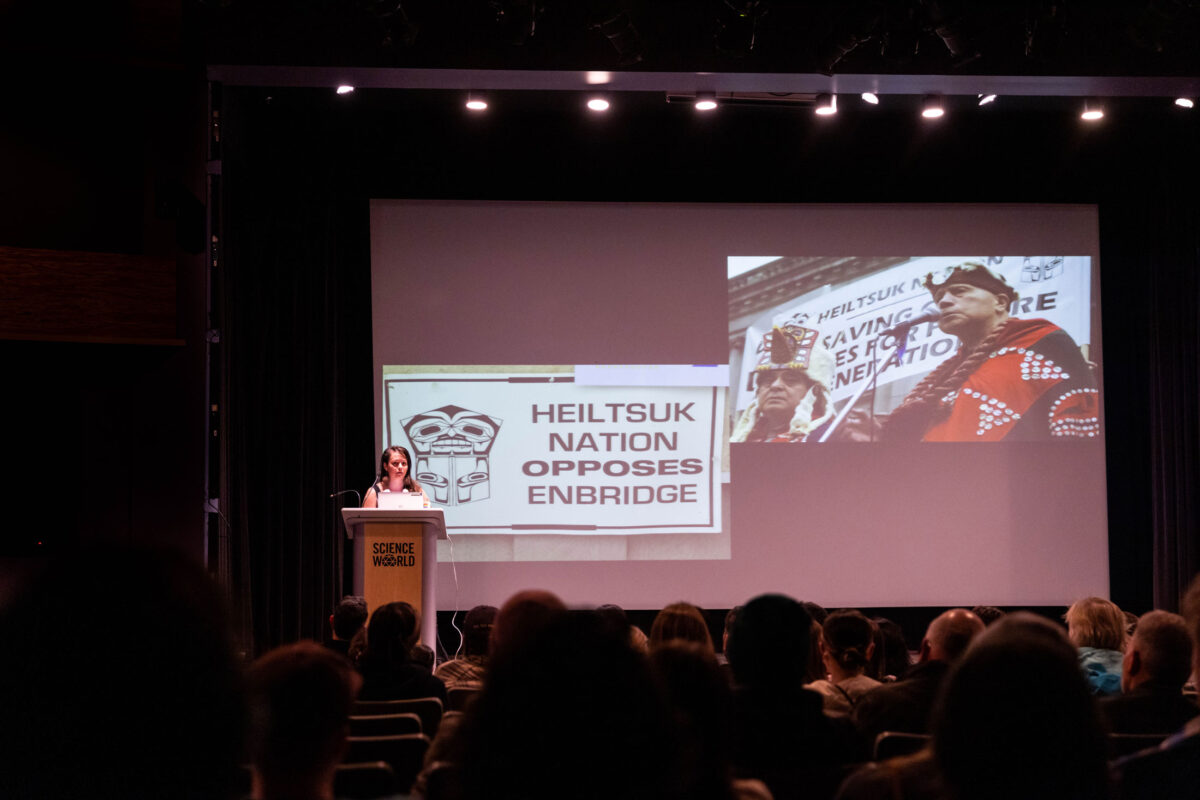 Jess Housty standing on stage with a slide of photos from the fight against Enbridge Northern Gateway Pipeline.