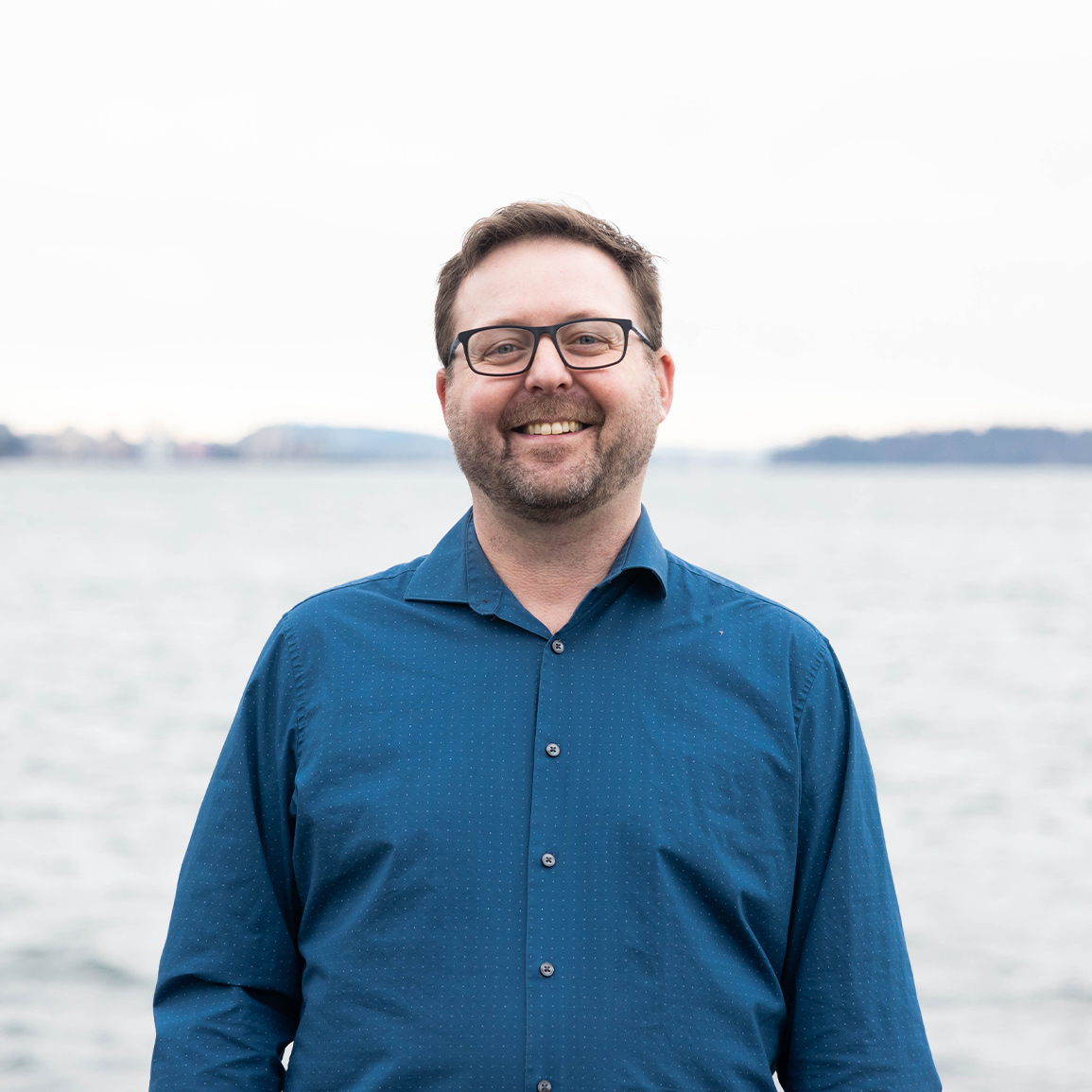Adam Warner standing in front of the ocean smiling, wearing a blue button down shirt.