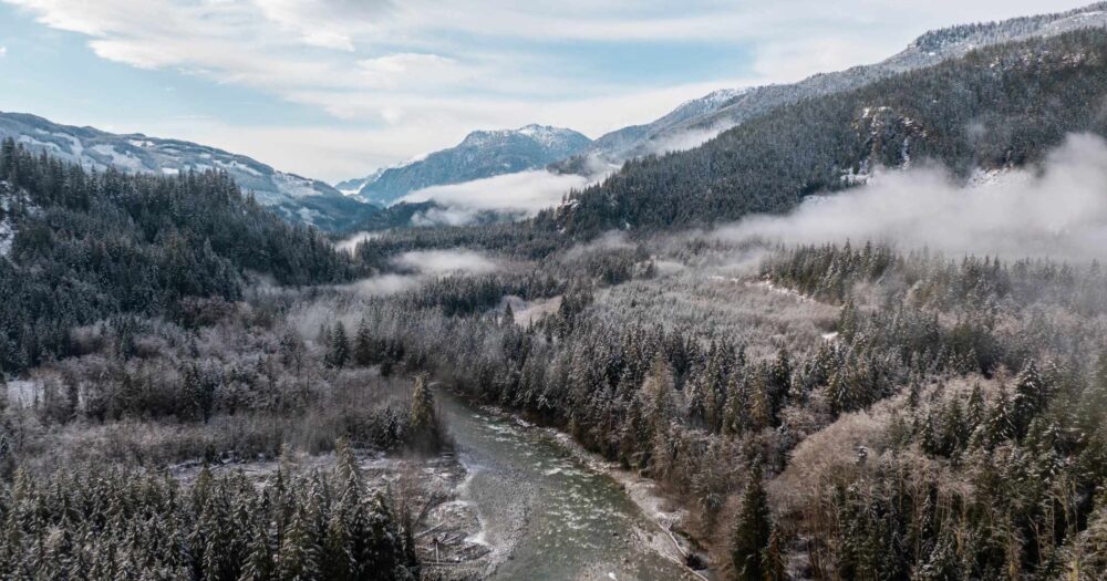 An aerial view of a river surrounded by snow covered forested mountains.