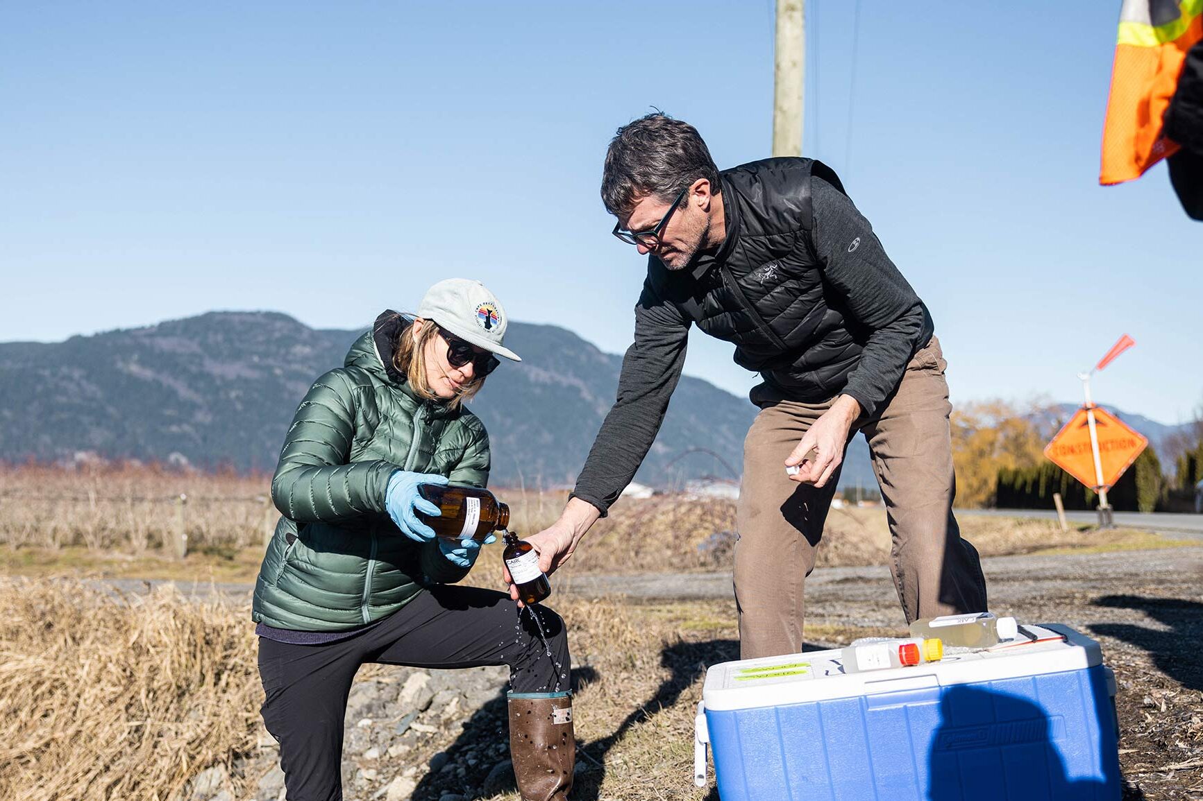 Two people working together to collect water samples in a farm field with the mountains in the background.