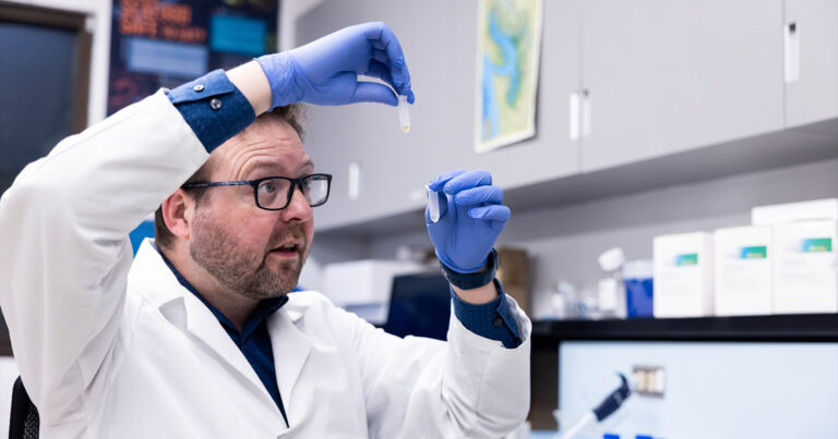 Research scientist Adam Warner holding two small beakers while wearing a lab coat in a lab.