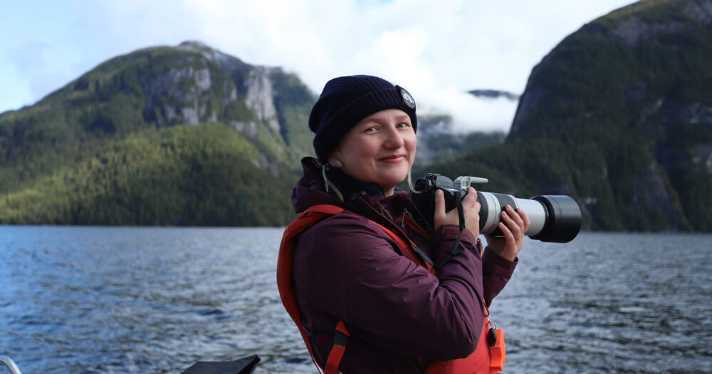 Erin Wall standing on a boat with beautiful mountains in the background holding a large camera.