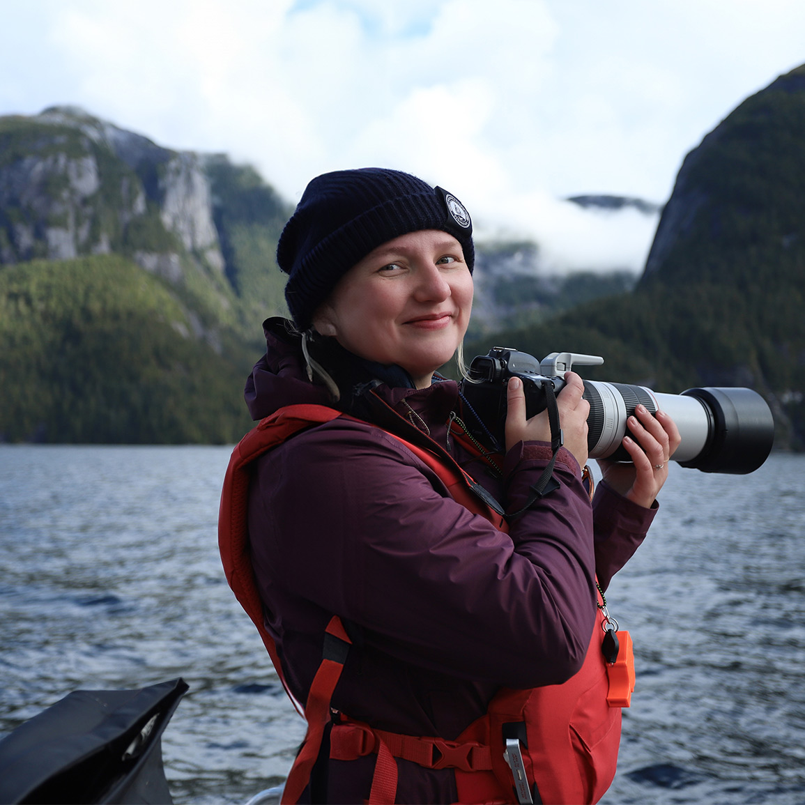 Erin Wall standing on a boat with beautiful mountains in the background holding a large camera.