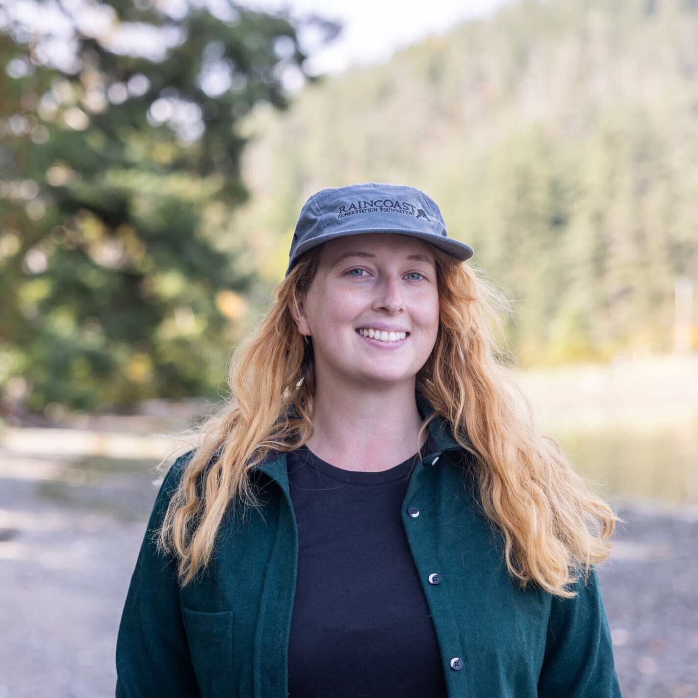 Sam has red hair and she is wearing a blue Raincoast hat, smiling while standing on a beach. 