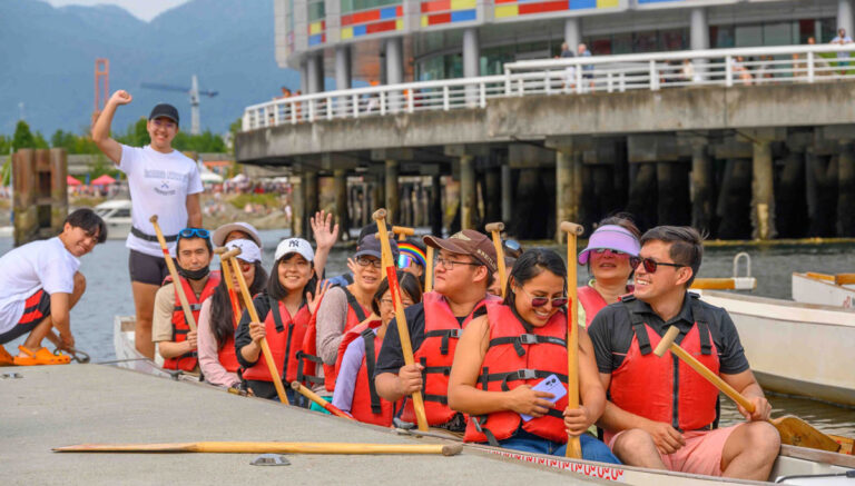 A team of people wave from a dragon boat outside of Creekside Community Centre and Dragon Zone Paddling.