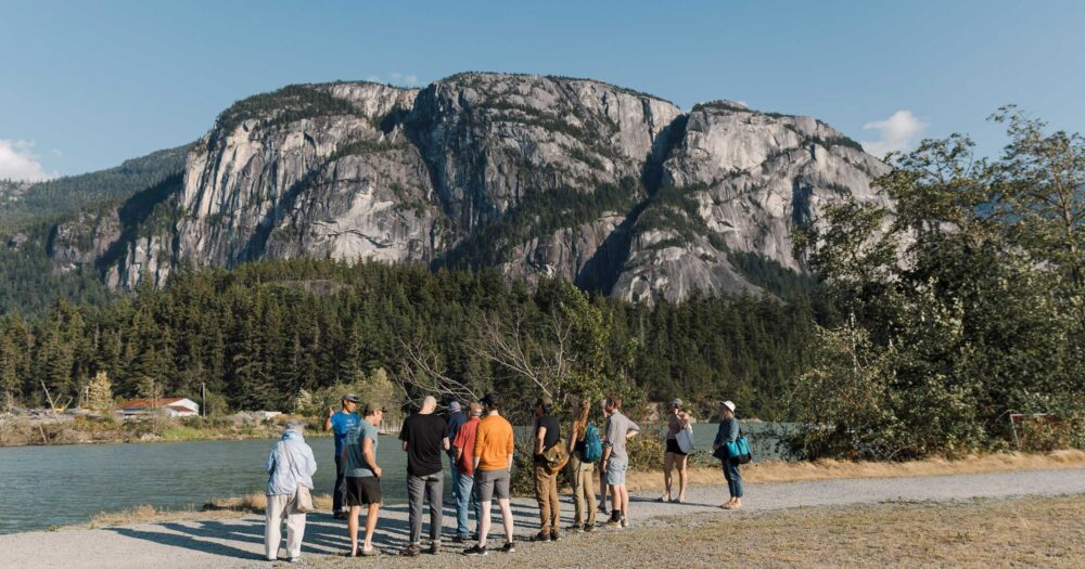 Group of people standing near the water with a big mountain in the background.