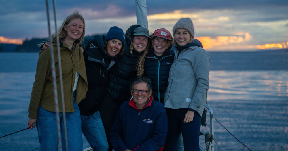 6 people pose together for a group photo onboard Achiever, with a backdrop of the sun setting over the ocean.