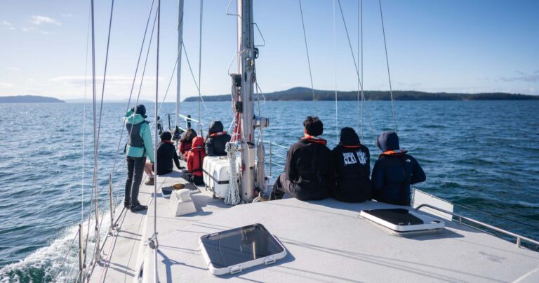 A swarm of youth sit on the bow of Achiever on a sunny day, with Pascale leading SSES curriculum.