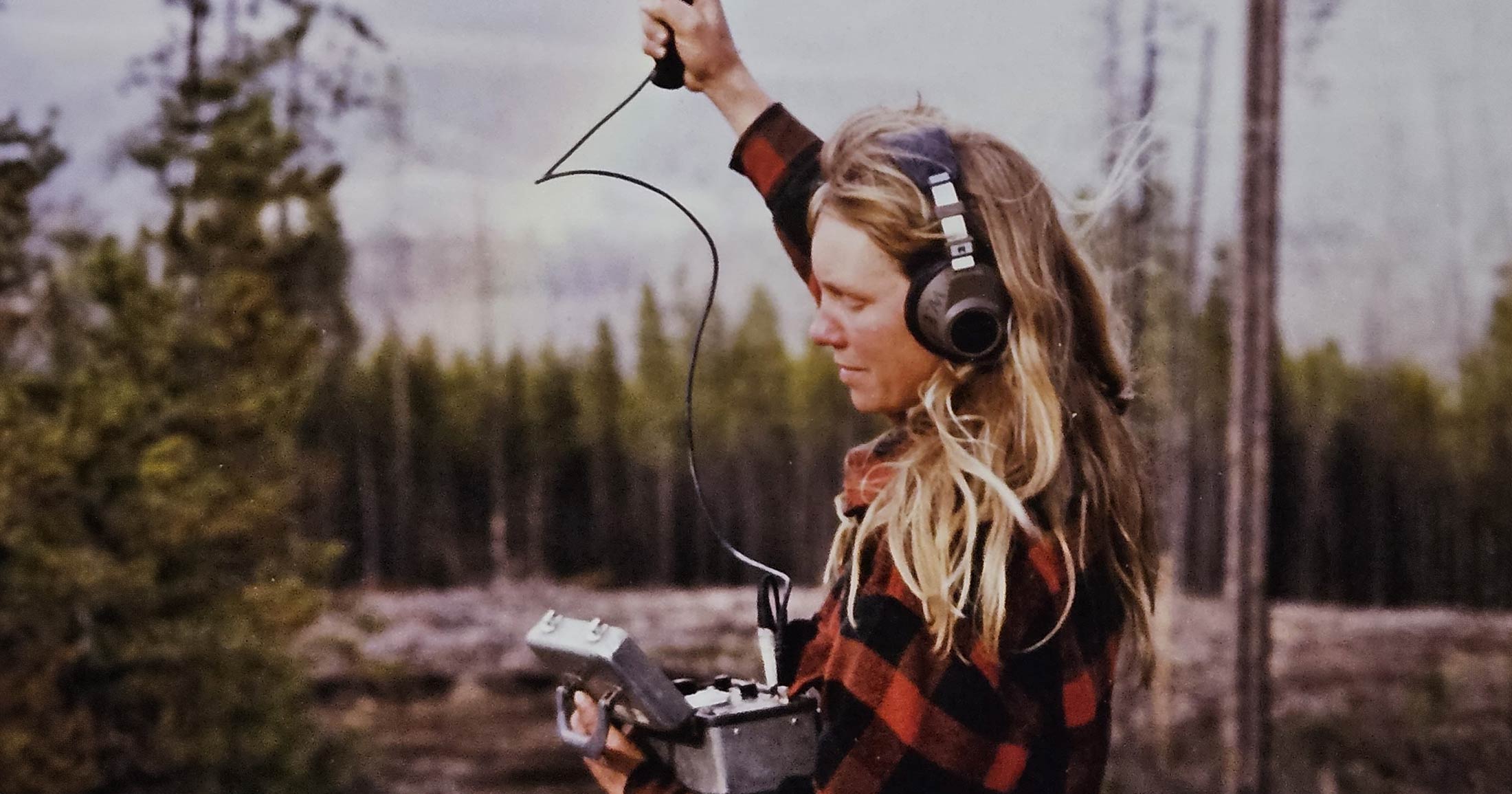 Diane Boyd stands in a cutblock holding a radio antenna up in the air while tracking wolves.