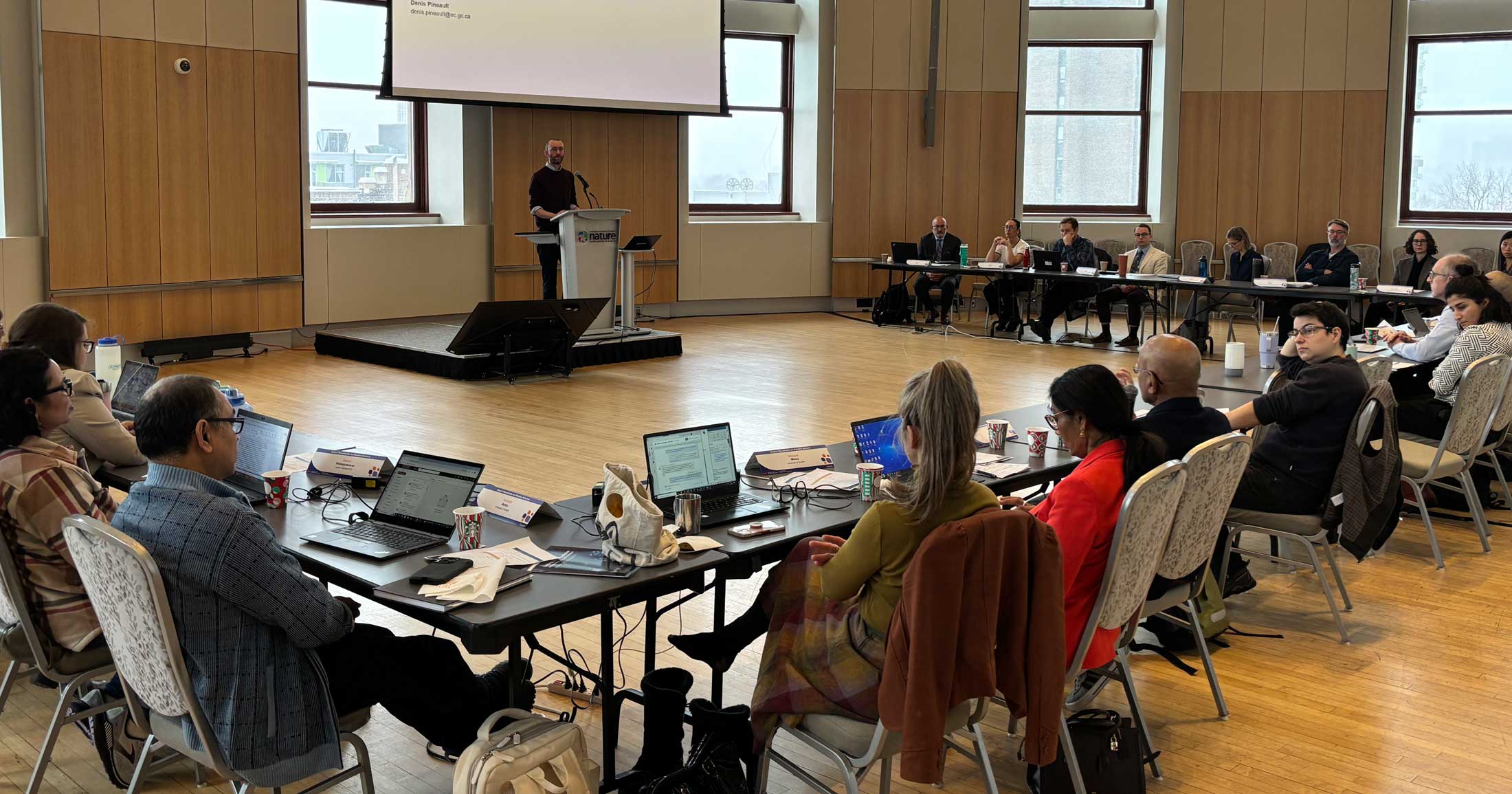 A panel of scientists sit around a large wide round table listening to a presenter.