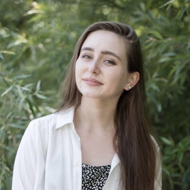 Nicole looks up to the left slightly, smiling happily, with long hair draping over her shoulder and surrounded by green foliage.