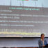 Kristen stands in front of a podium with a microphone presenting a power-point that reads "Post wildfire logging"