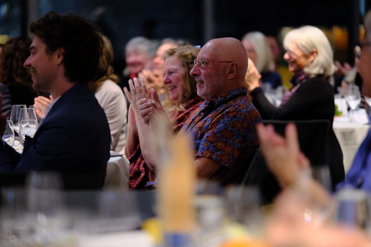 A smiling and clapping audience looks towards speaker standing out of frame.