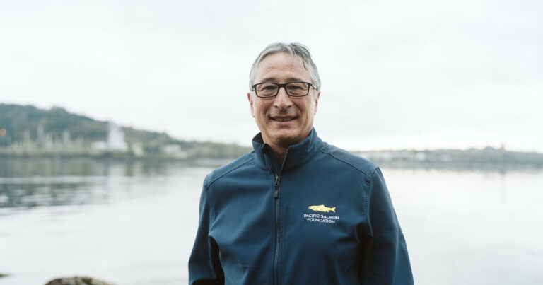 A man smiles at the camera, standing in front of a calm body of water, wearing a deep blue jacket.