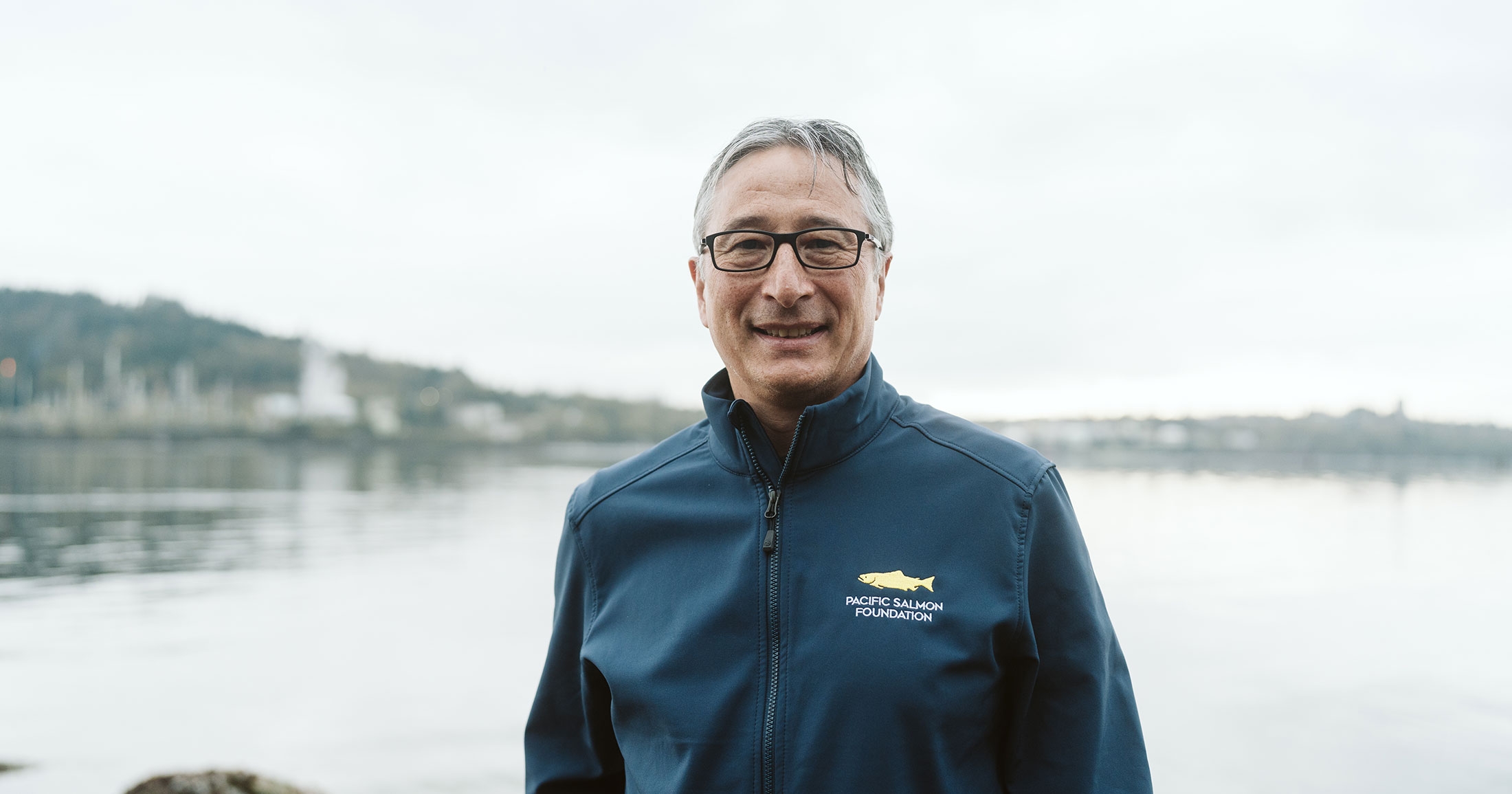 A man smiles at the camera, standing in front of a calm body of water, wearing a deep blue jacket.