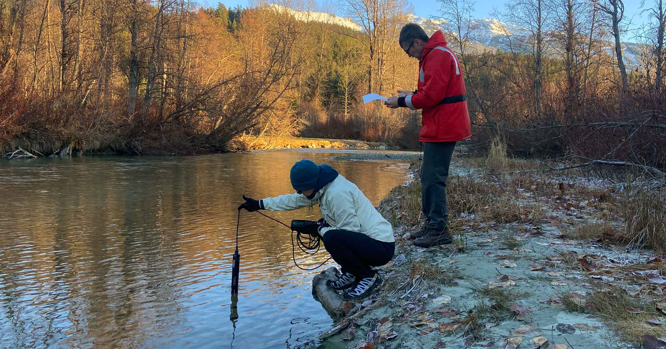 Two scientists stand at the edge of a body of water, one standing up, wearing a red raincoat and writing notes, the other crouching and deploying a tool into the water on a long cord.