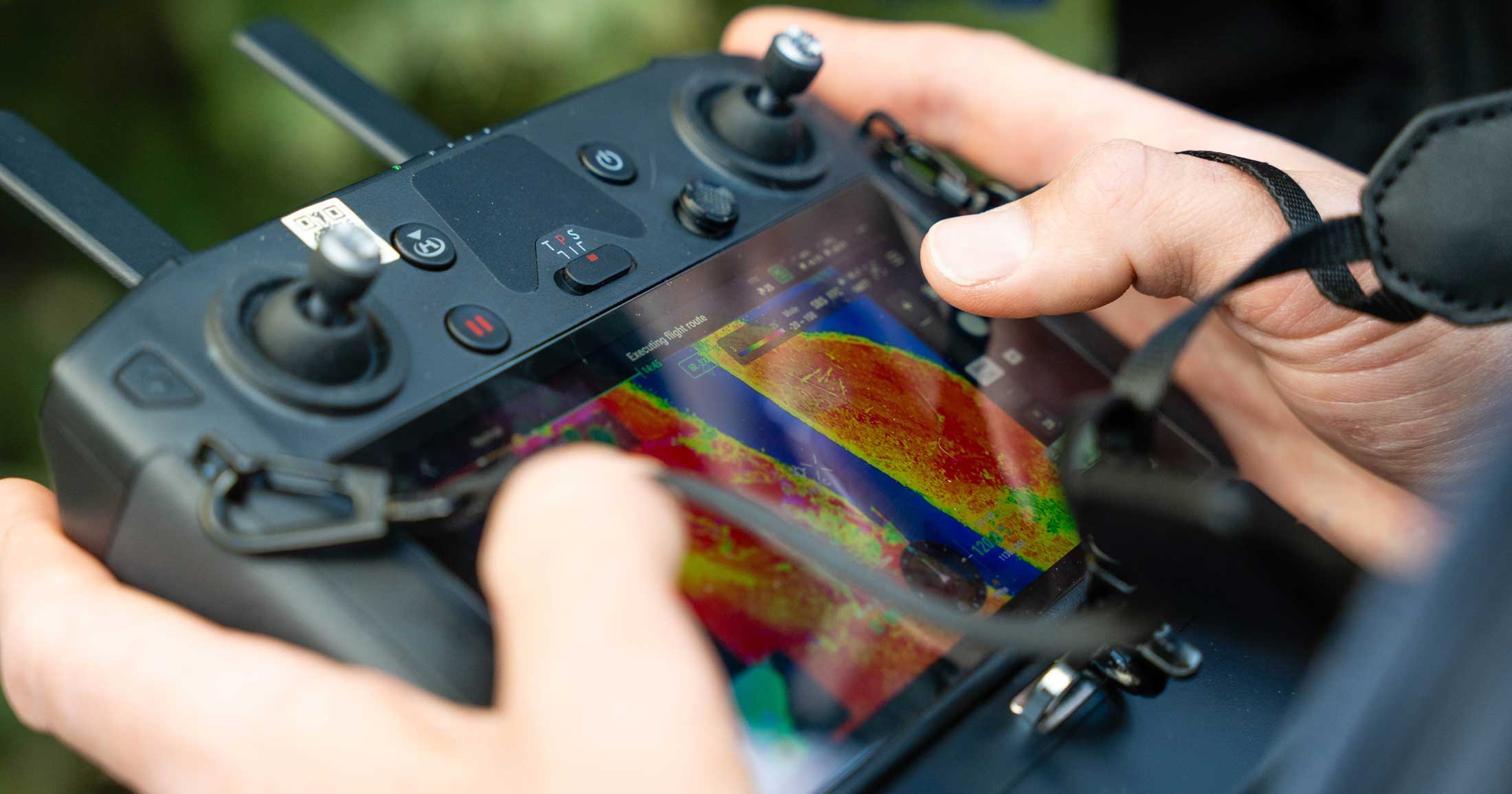 Two hands hold a drone controller, the display showing bright orange and yellow thermal areas.