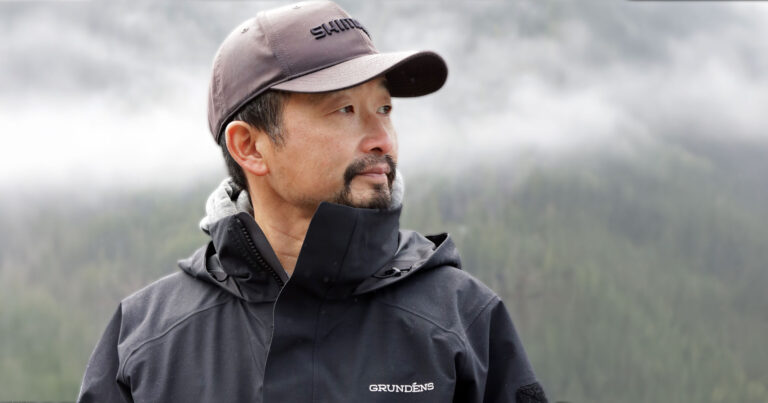 Rodney Hsu looks out over the water with mist and clouds and mountains looming behind him.