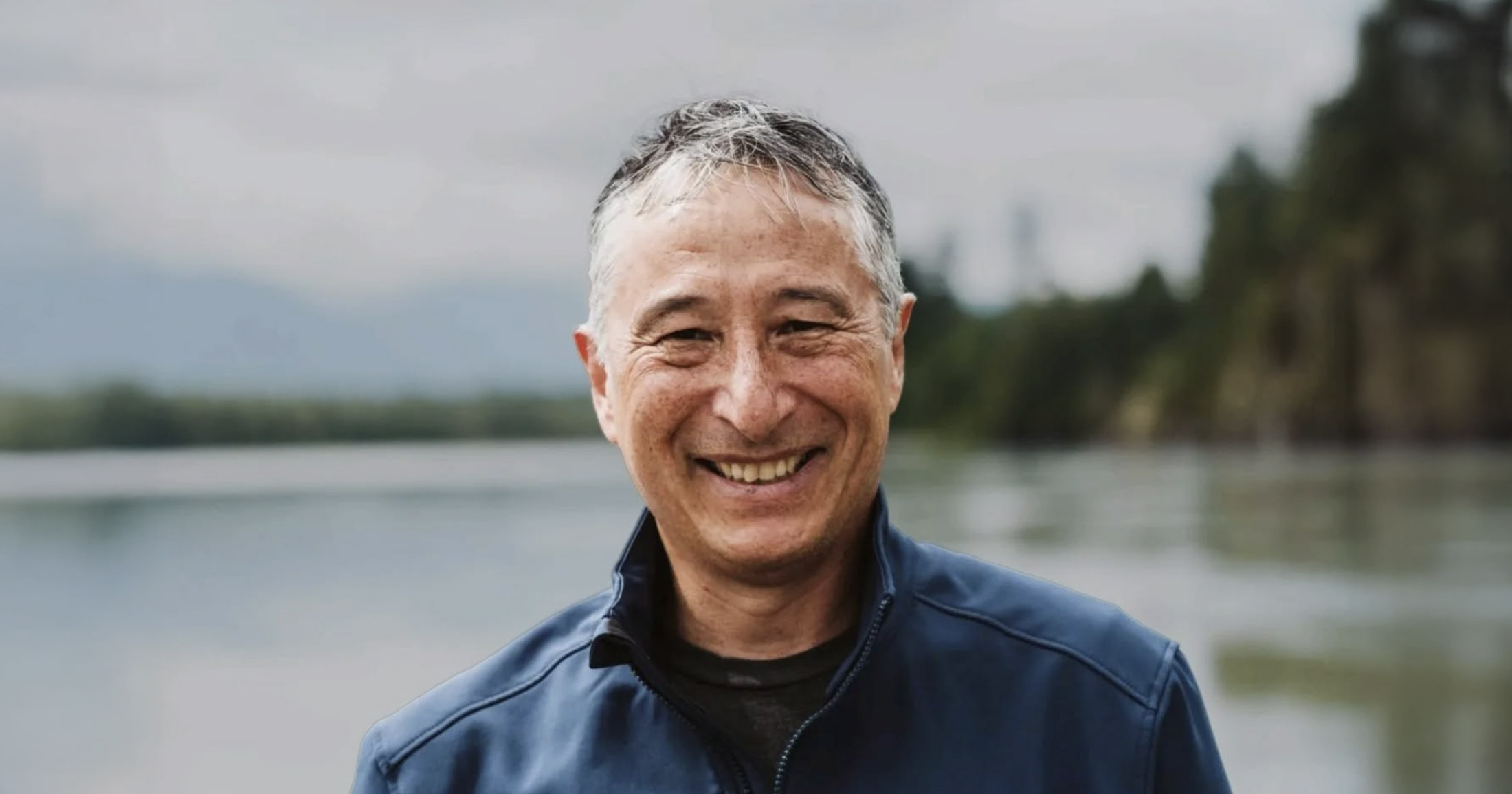 Jason Hwang stands in front of the water wearing his Pacific Salmon Foundation jacket, smiling widely without his glasses.