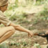 A young Jane Goodall extends her right hand towards a baby chimp, crouched down in a remote jungle scene.