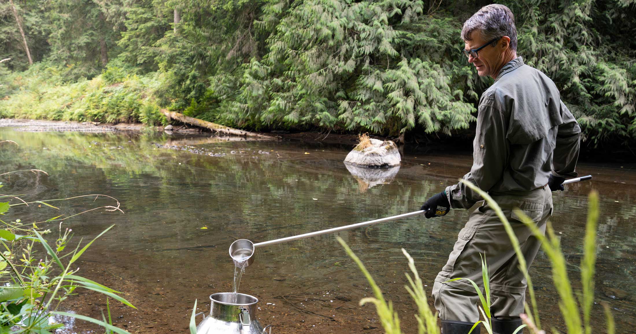 A scientist stands in field clothes by a stream, seen through foliage as he collects a water sample by using a long silver rod that has a cup on the end. He collects water from the stream and dumps it into a large silver jug.