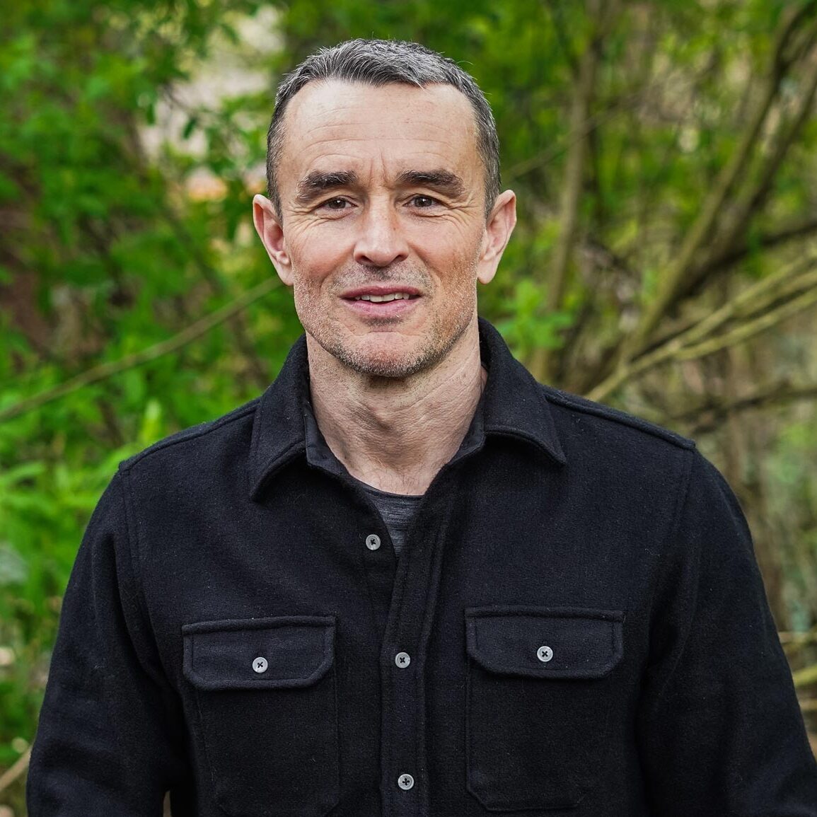 Chris Darimont stands in the woods with a green backdrop, wearing a dark heavy button up shirt and collar.