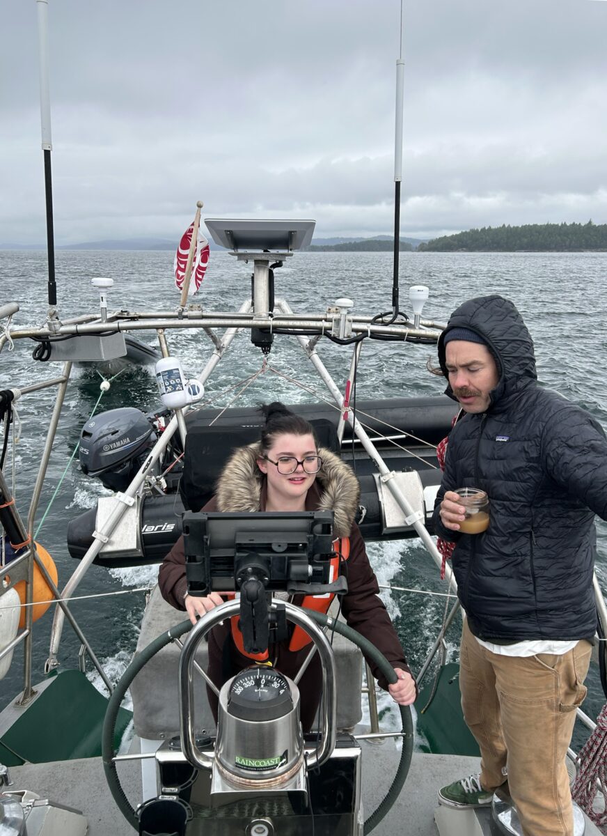 A teenager at the helm of a sailboat while the captain looks on.
