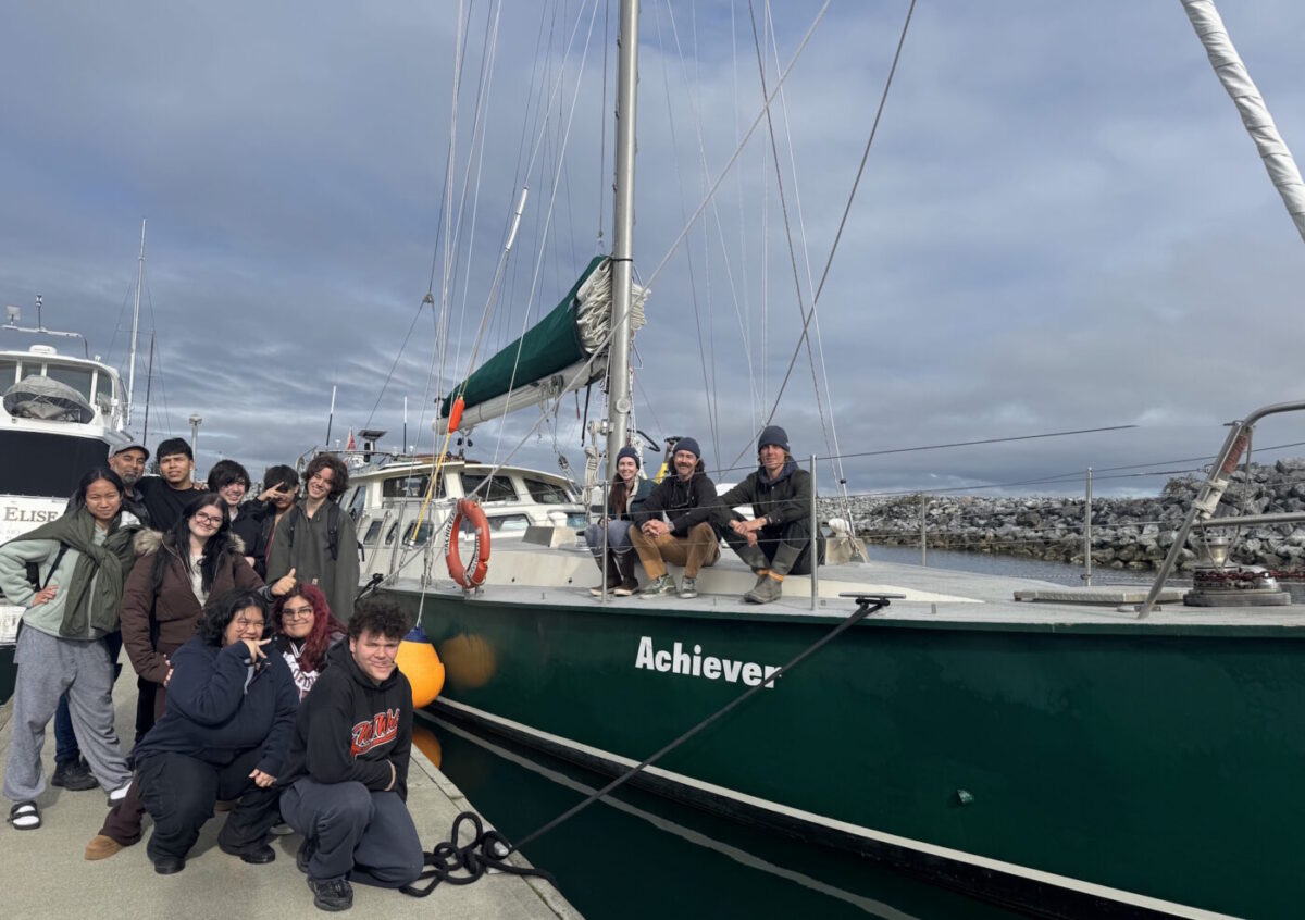 A group of youth participants pose next to the sailboat Achiever and the crew