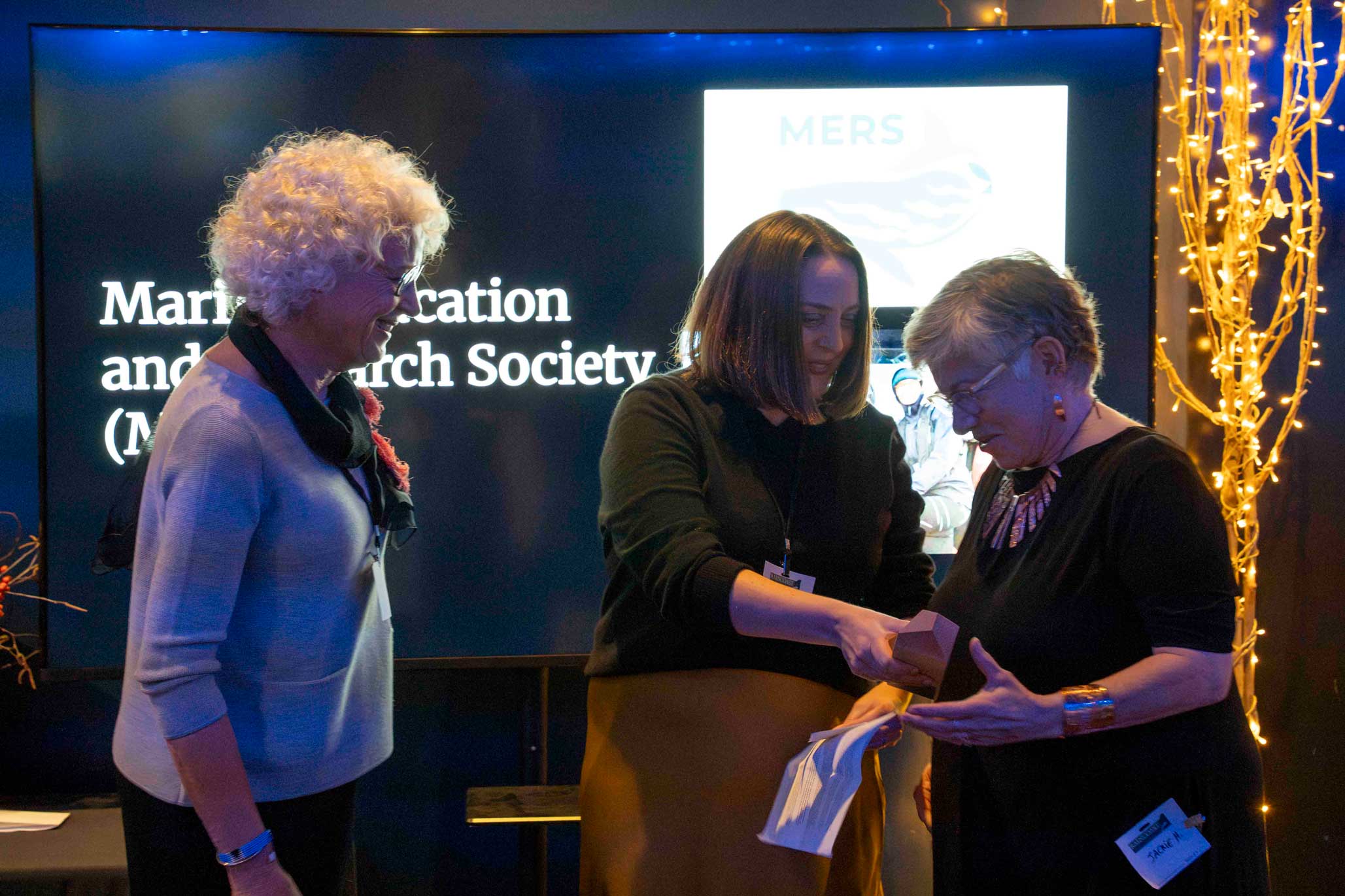 Three women stand in front of a bright screen, smiling at one another and the middle individual handing an award to the individual on the right, the other smiling fondly at the two of them.