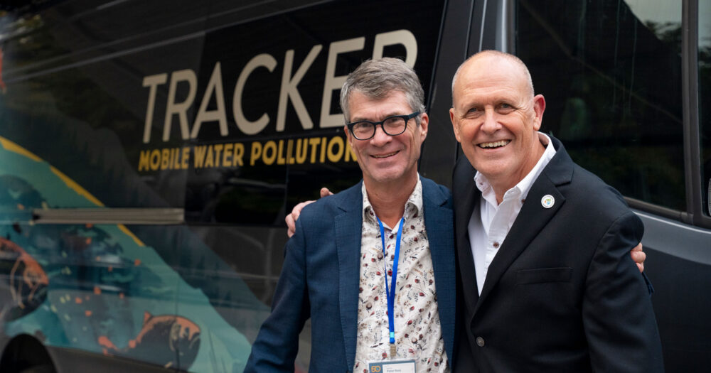 Dr. Peter Ross and Clint Wright stand arm in arm smiling at the camera, with a sprinter van that has been converted into a mobile water pollution lab visible behind them.