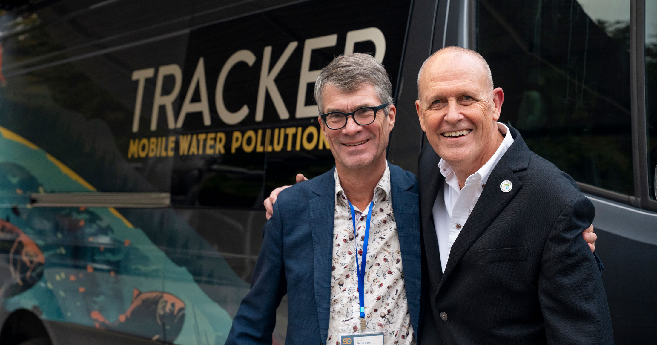 Dr. Peter Ross and Clint Wright stand arm in arm smiling at the camera, with a sprinter van that has been converted into a mobile water pollution lab visible behind them.