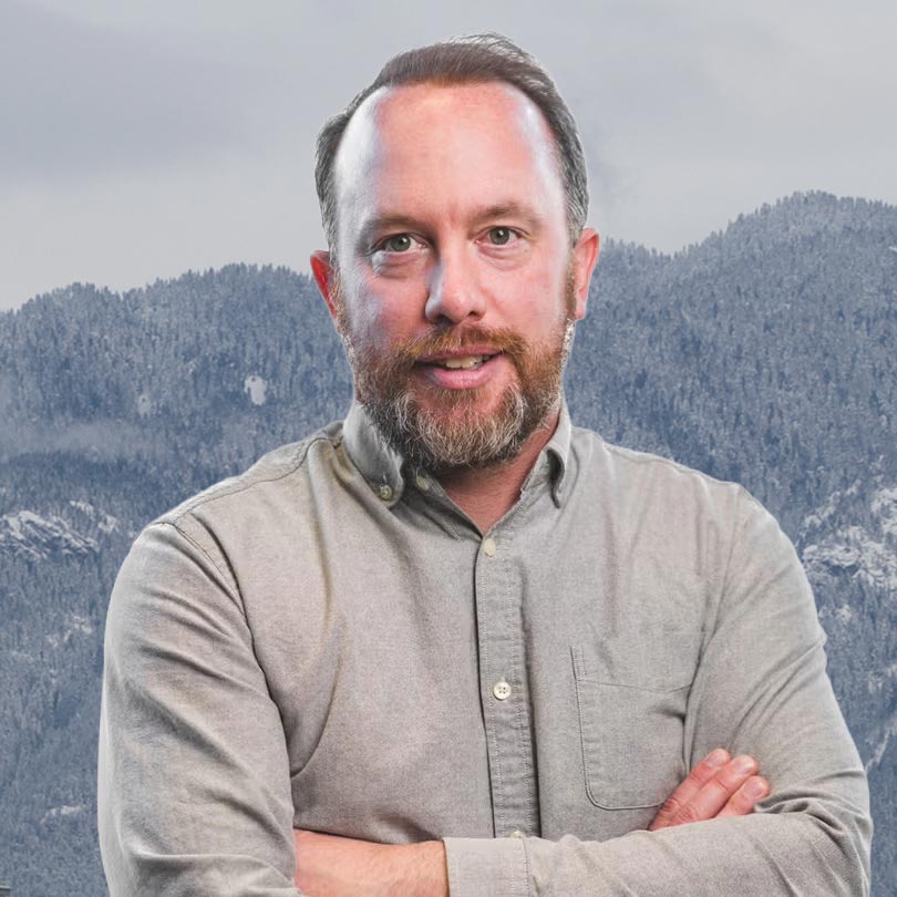 Jeffery Young stands with his arms crossed in a sharp collared shirt with snow mountains looming behind him.